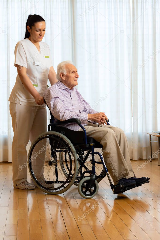 Full length portrait of old handicapped man sitting in wheelchair. Young female nurse standing next to senior patient in living room.
