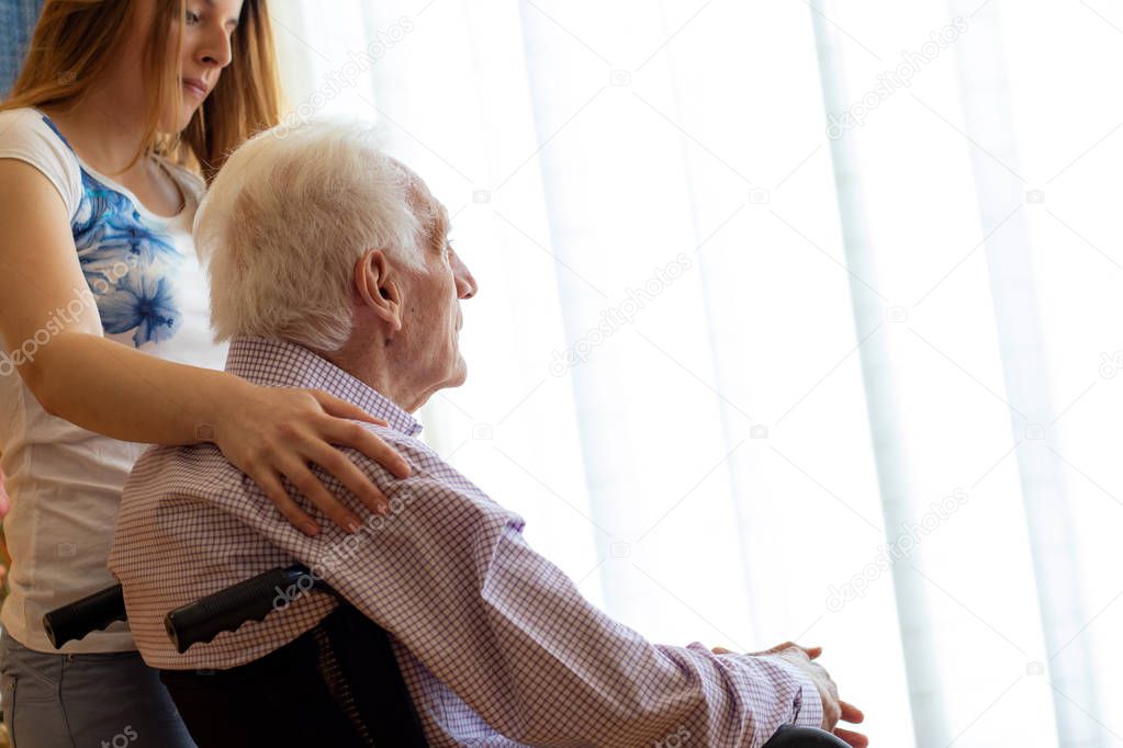 Close up side view portrait of old man in wheelchair staring at window. Young female caregiver embracing man with hand on shoulder.