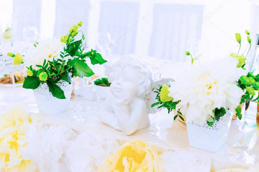 a statue of a little angel at the tables for newlyweds among a vase with beautiful flowers and a tablecloth decorated with flowers from the fabric