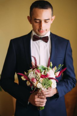 Beyaz gömlekli ve papyonlu damat, arka planda bir düğün buketi tutuyor.