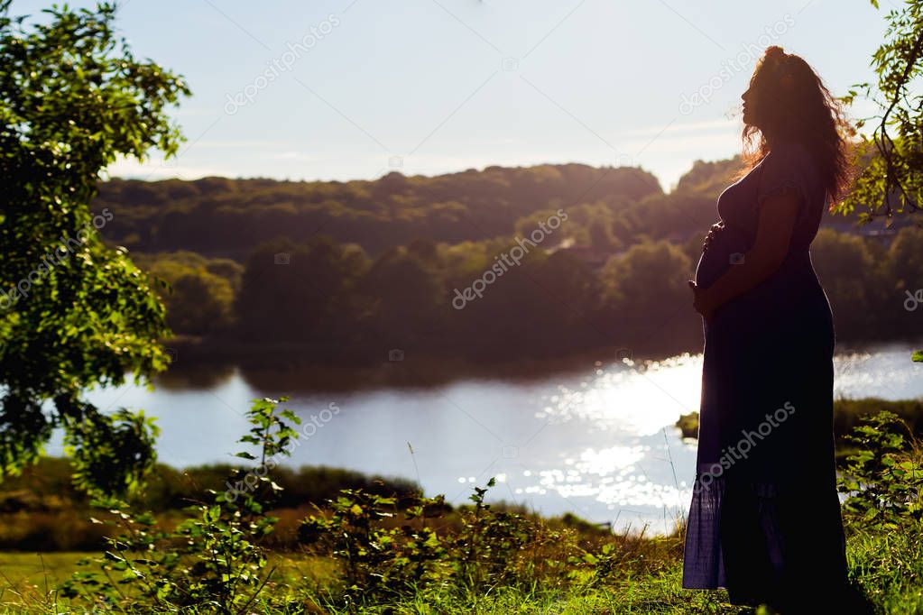 a pregnant woman in a beautiful dress is standing on the background of the picturesque lake
