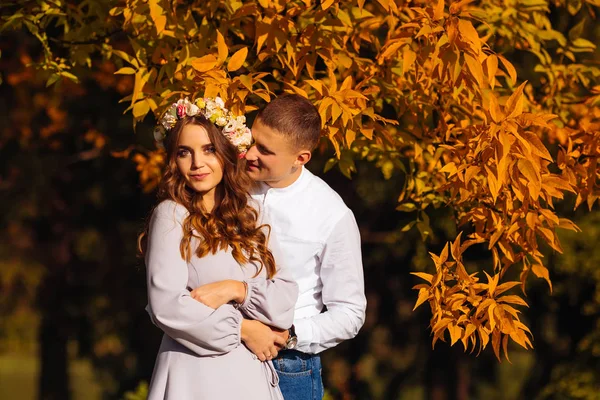 頭に花輪を持つかわいい女性がカメラのレンズを見て 彼女の夫は黄色の秋の木々 を背景に彼女を抱擁 — ストック写真