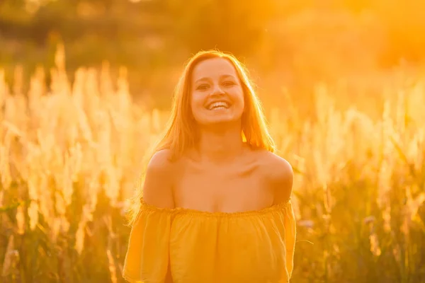 美丽的笑容女孩在黄色的礼服与裸露的肩膀在夕阳和高草的背景下 — 图库照片