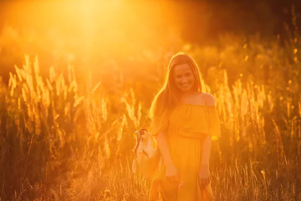 女孩在一个时髦的礼服和背包在她的肩膀看着相机镜头对背景的日落 — 图库照片