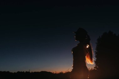 Yapay ışığın parladığı bir şapkayı tamir eden bir kızın karanlık silueti. Kız akşam gökyüzünün arka planında ve ağaçların siluetinde duruyor.