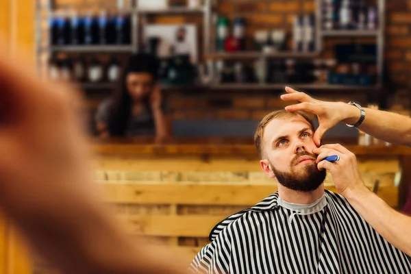 A man in a barber studio with a long beard sits in a chair and the hairdresser cuts beard on the background of the reception
