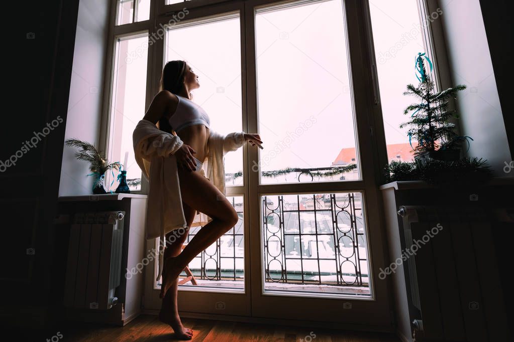 Beautiful sexy girl in white dressing gown standing near the window with in luxury modern apartment, looking at cityscape after waking up in the morning, relaxing. Perfect morning.