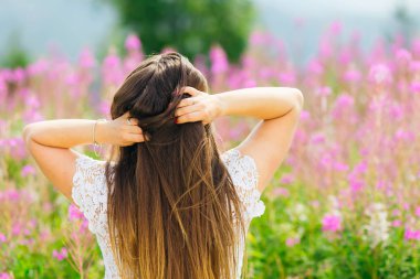 Dantel bir elbise içinde gelin kameranın arkasında duruyor ve yabani çiçeklerin arka planına karşı saç ellerini tutar.