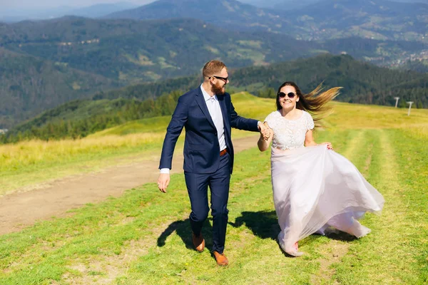 新郎穿着时髦的西装 留着胡子和太阳镜 手牵着新娘 他们在山顶上的草地上奔跑 — 图库照片