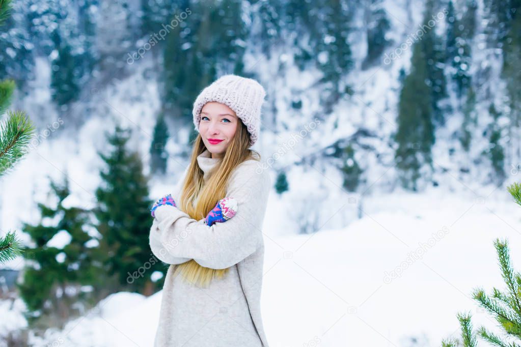 girl in a warm sweater and hat embracing herself against the background of snow-covered mountains and forests
