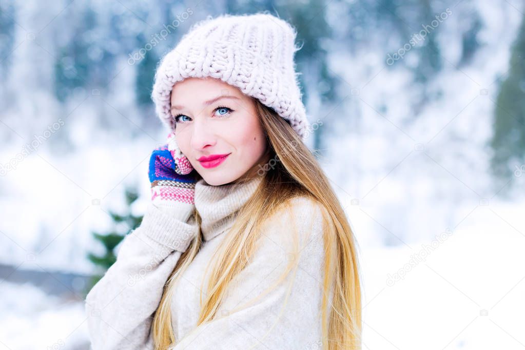 Close-up of a girl's face in a sweater and a hat that looks in the camera lens on the background of snow and wood