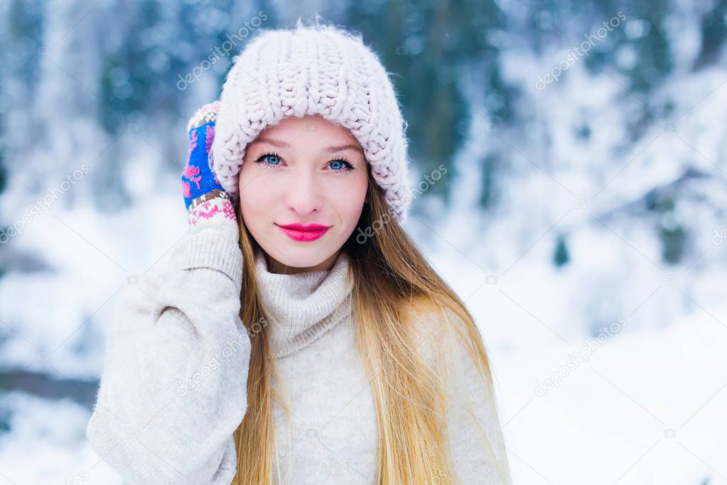 The girl holds her hand on the hat and looks at the camera lens against the background of snow and forests
