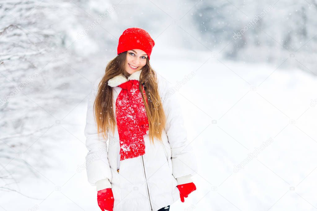 girl in a white winter jacket and red hat with a scarf looking in the camera lens and smiles on the background of the trees and the winter park