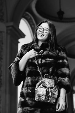 Bir kız şık kış giysileri ve bir sırt çantası siyah ve beyaz fotoğraf üzerinde building yakınındaki uzak ve sevimli gülümsüyor görünüyor