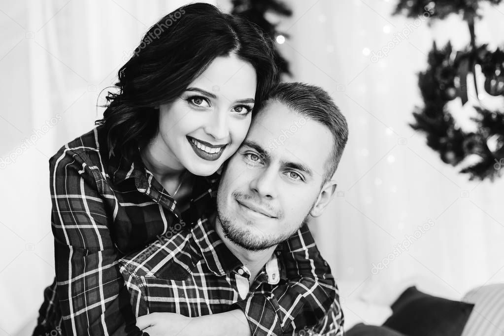 close-up couple who are looking into the camera lens and smiling on a black and white photo. they are sitting on a bed with Christmas decorations