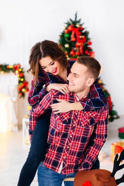 The girl jumped on the back of the guy and they smile against the background of a christmas tree and a fireplace