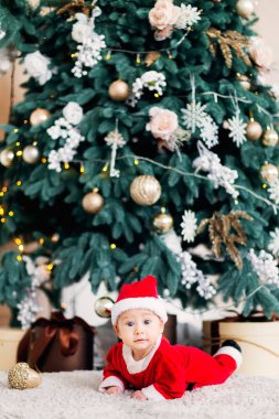 bir Noel Baba kostüm bir mideyle yalan Noel ağacı yakınındaki ve sunar ve kamera lensi görünüyor bebek