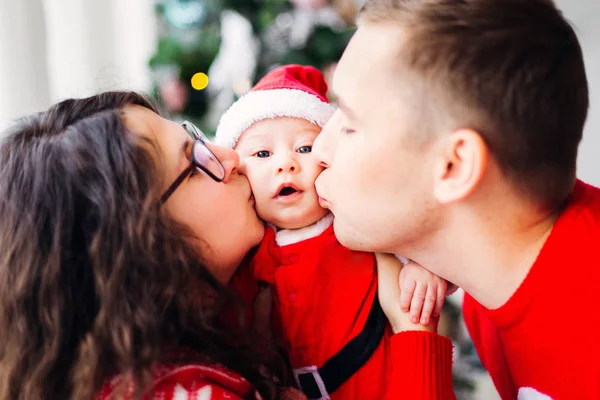 dad and mom kissing son\'s cheeks on both sides of him and the baby in a Santa Claus costume looks in the camera lens