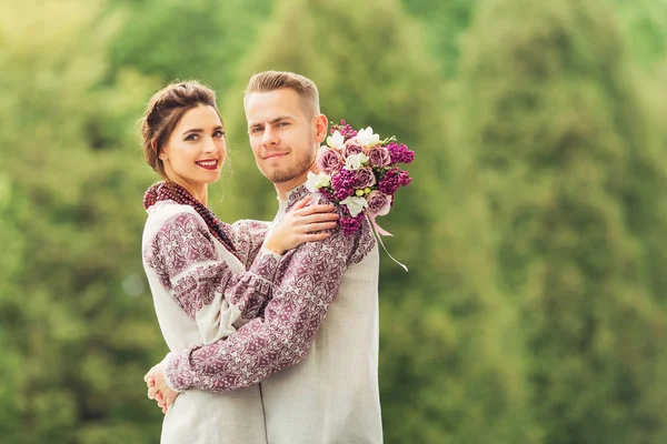 穿着节日服装的新婚夫妇互相拥抱 看着镜头 微笑着在树的背景上 新娘拿着美丽的婚礼花束 — 图库照片