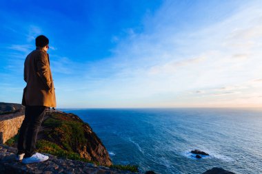şık giysiler giyen bir kayalık sahil kamerada duran ve okyanus ve gün batımı hayran