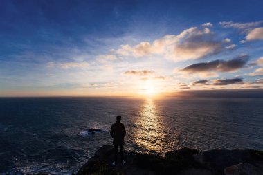 şık giysiler giyen bir kayalık sahil kamerada standları ve günbatımı okyanus bakar