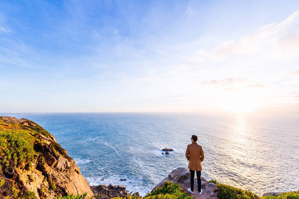 man in stylish clothes stands back to the camera on a rocky shore and looks at the sunset and the ocean