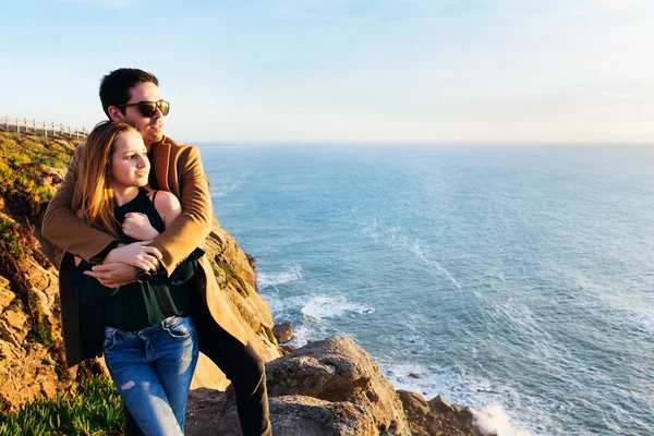 guy in a coat embraces a girl in a blouse and they admire the evening ocean from a high rocky shore