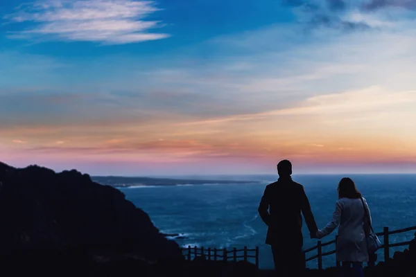 Couple silhouettes at dusk that hold hands and stand back to the camera on the background of rocky hills and the ocean