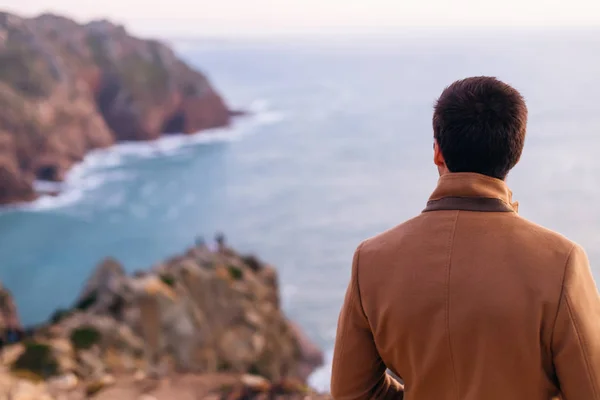 A close-up of a guy standing back to the camera in a beige coat on the rocky coast of the ocean