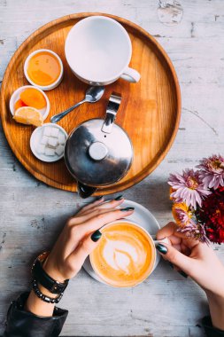 bir fincan kahve bir köpük ve kızın elleri ile en iyi görünümü. boş bir bardak, bir metal su ısıtıcısı ve şeker bal ve meyve dilimleri ile ahşap tepsi