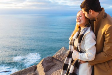 Adam arka yüzünde kızdan hugs ve onun gözleri ve gülümsüyor Açıkçası üzerinde kayalık sahil akşam okyanus manzarası ile kapatır