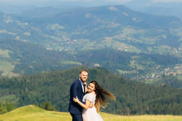 the newlyweds smile and look at the camera lens against the backdrop of green mountains