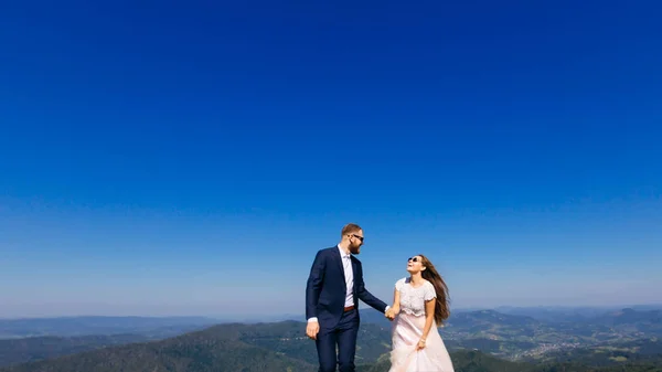 新郎的胡子穿着时尚的西装 地平线和天空的背景下 穿着白色的衣服 戴着太阳镜 握着新娘的手 — 图库照片