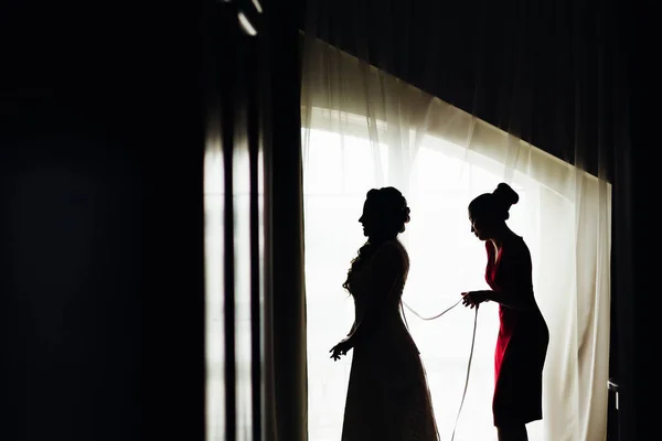 結婚式のコルセットのひもを結んで 窓の近くの格好をした花嫁および彼女の友人のプロファイル — ストック写真
