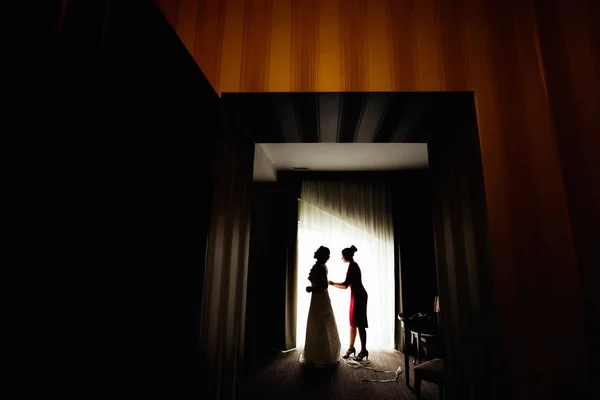 新娘的费用在靠近窗户的黑暗房间里 新娘的女朋友帮忙在婚纱上系上紧身胸衣 — 图库照片