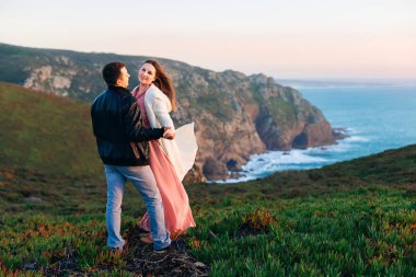Adam ayakta geri kamera ve tutar ellerini, kız kameraya bakıyor. kayalık sahil ve akşam okyanus