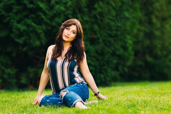 Een mooi meisje zit op het gras in het Park. Kijk naar camera — Stockfoto