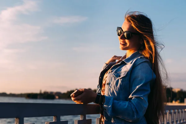 Стильная девушка в солнечных очках возле забора у озера. девушка улыбается и — стоковое фото