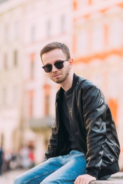 Siyah deri ceketli ve güneş gözlüklü sakallı adam ona bakıyor.