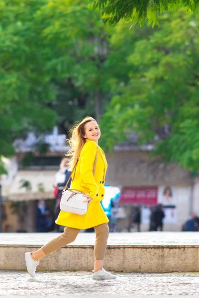 Menina em um casaco amarelo e bolsa sorri e caminha. rua da cidade — Fotografia de Stock