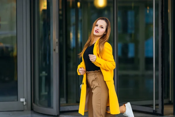 Девушка в стильном желтом пальто смотрит в камеру и поднимает — стоковое фото