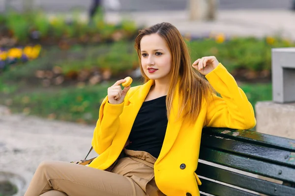 Beautiful girl sitting on a bench and holding yellow macaroon in — Stock Photo, Image