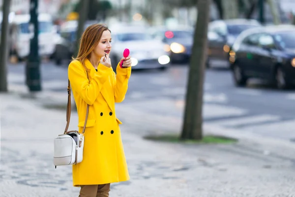 Ragazza in un cappotto giallo con una borsetta che applica il rossetto sulla m — Foto Stock