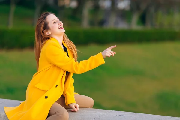 La ragazza è seduta in un cappotto giallo e mostra qualcosa. strada — Foto Stock