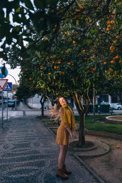 Girl in a dress on the sidewalk waving her hair. trees with oran — Stock Photo, Image