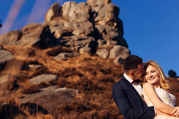 Ο γαμπρός αγκαλιάζει τη νύφη και χαμογελούν στο φόντο του βουνού — Φωτογραφία Αρχείου