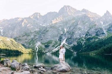 Birbirine aşık çiftler taşın üstünde sarılıyor. Kayalık dağlar, göl ve f