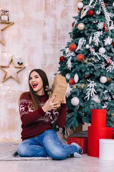 Jovem segurando uma caixa de presente e sentado perto de uma árvore de Natal — Fotografia de Stock