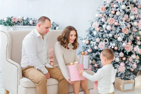 mom and dad sit on the sofa near the Christmas tree and give a g