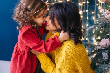 Küçük kız, Noel 'in yanında sarı kazaklı annesine sarılıyor.